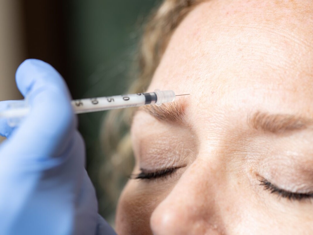 A gloved hand holds a needle to inject something into a woman's forehead, suggesting a Botox injection