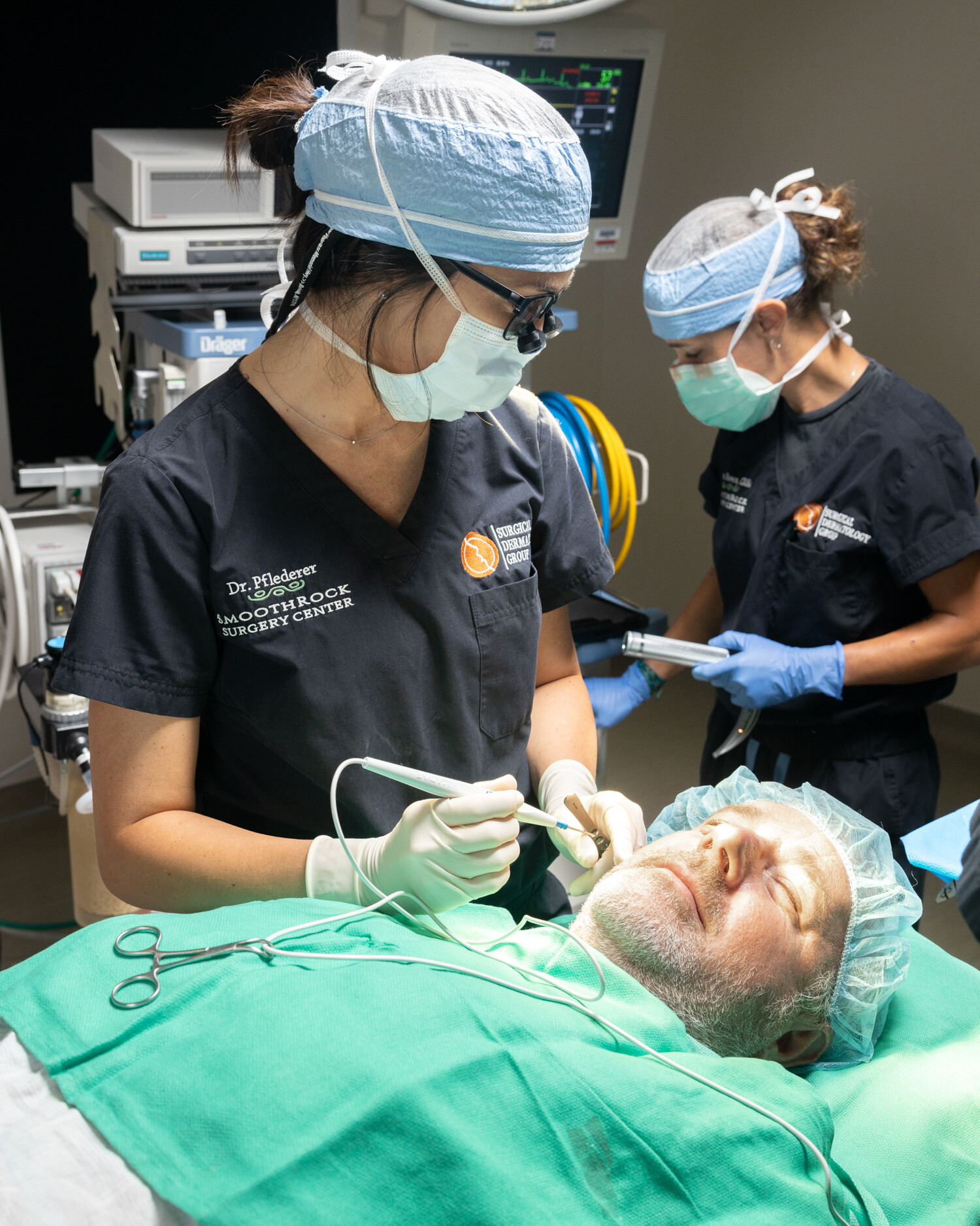 A doctor does a procedure on a patient with a medical provider in the background at Surgical Dermatology Group